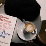 A café setting with a latte, book by Gloria Steinem, and cozy hat on a table.
