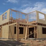 A two-story wooden house frame is under construction on a clear, sunny day.