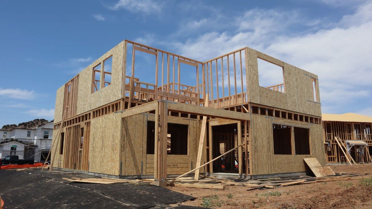 A two-story wooden house frame is under construction on a clear, sunny day.