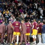 Basketball team huddles on court during a game timeout with audience in background.