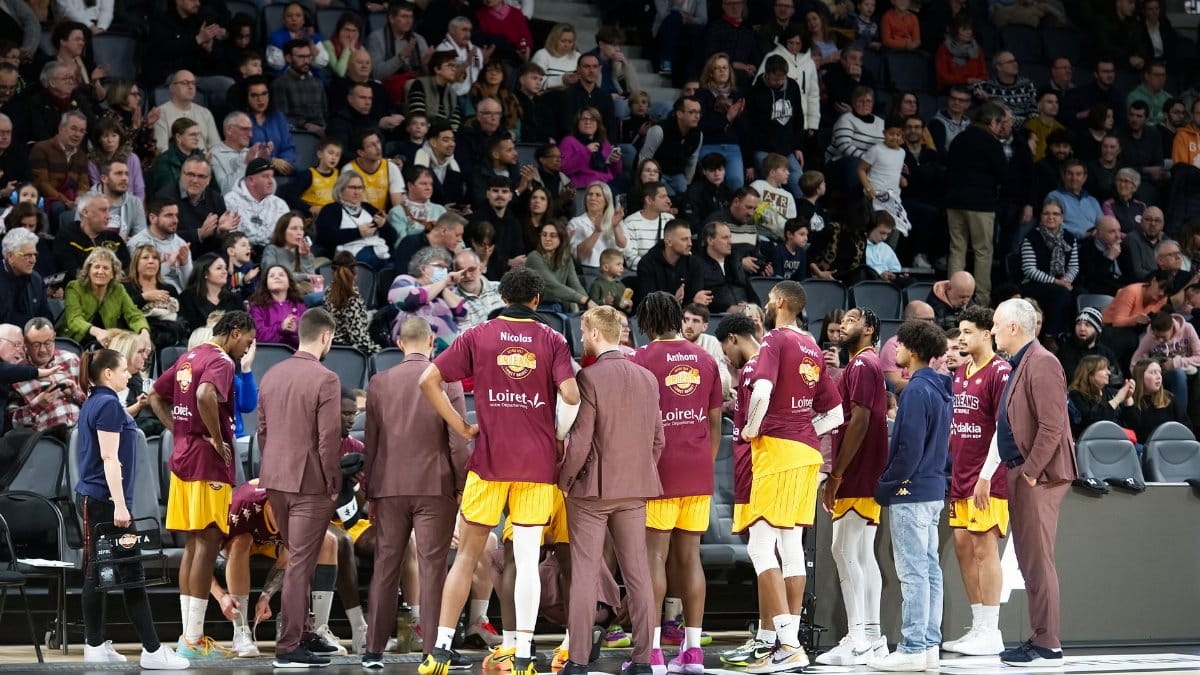 Basketball team huddles on court during a game timeout with audience in background.