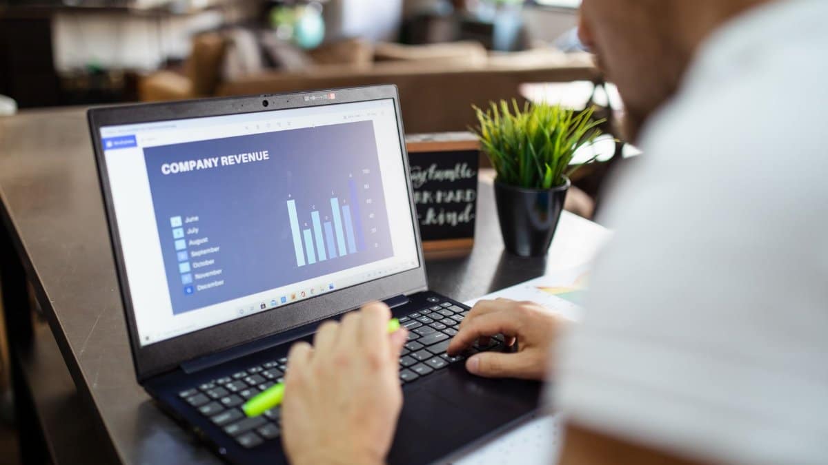Man using a laptop to analyze company revenue data with graph charts in office setting.