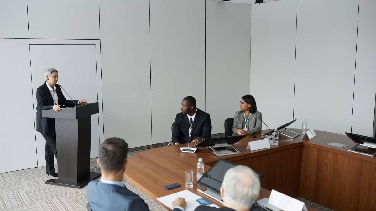 Diverse group of professionals in a formal meeting setting with a speaker at the podium.
