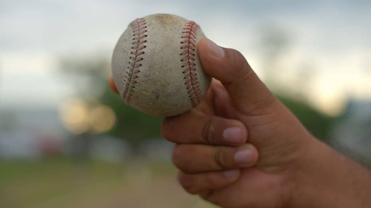 Detailed close-up of a hand holding a baseball outdoors with a blurred background, showcasing grip and texture.