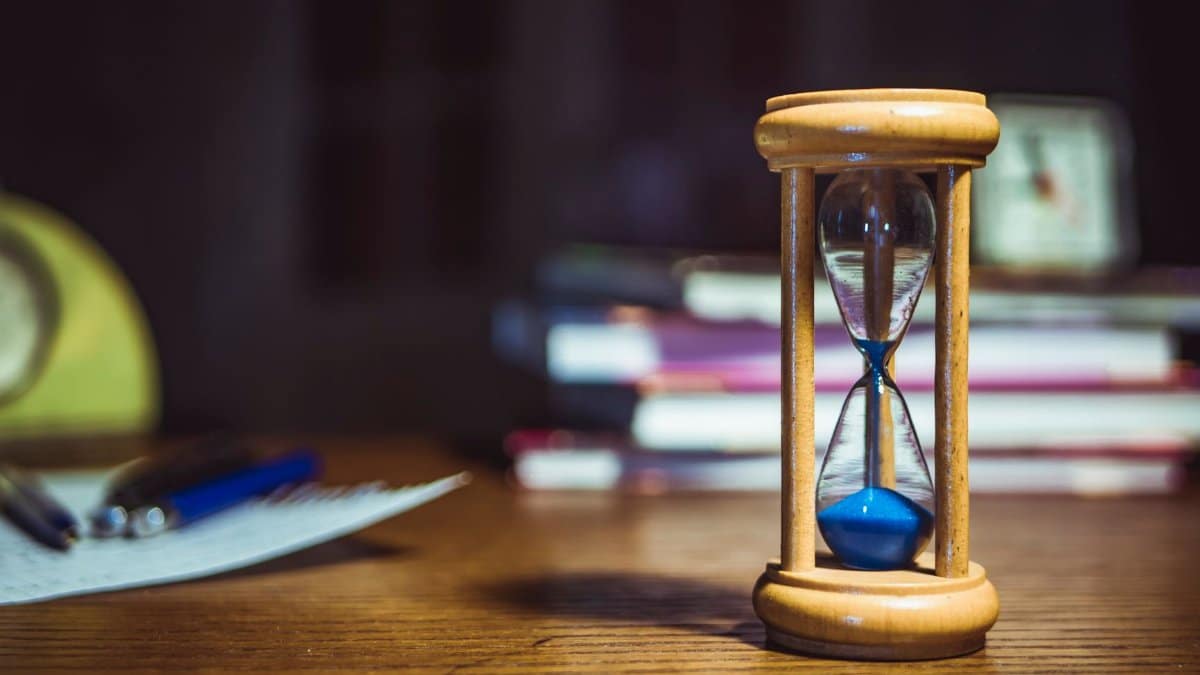 Close-up of a wooden hourglass on a desk, alongside books and pens, suggesting focused study or time management.