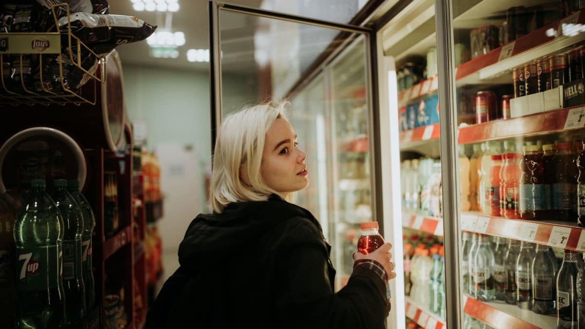 Blonde woman selecting a beverage from a refrigerated shelf at a convenience store.