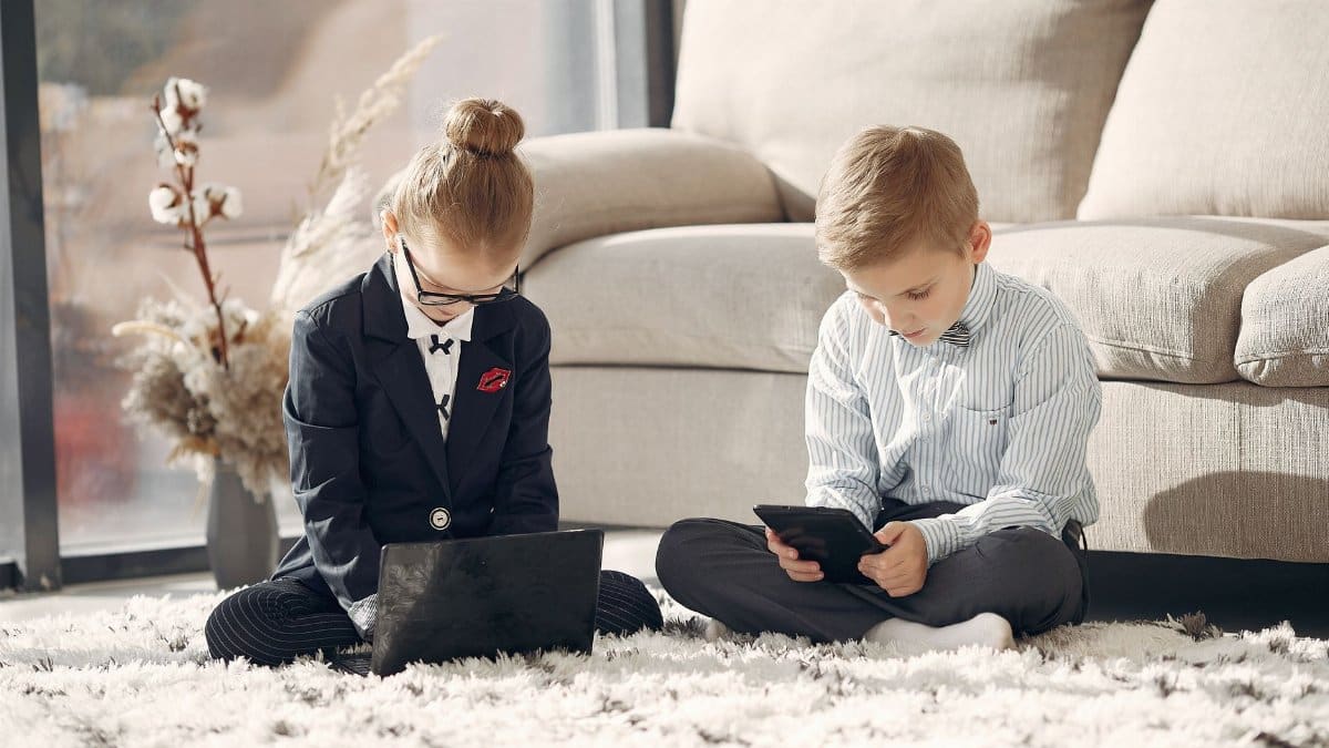 Serious kids in formal wear and eyeglasses sitting on carpet surfing table and laptop while working remotely in living room