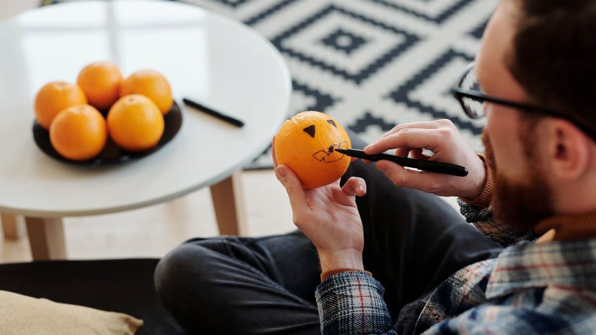 A man decorates an orange with a marker, creating a Halloween-style design indoors.
