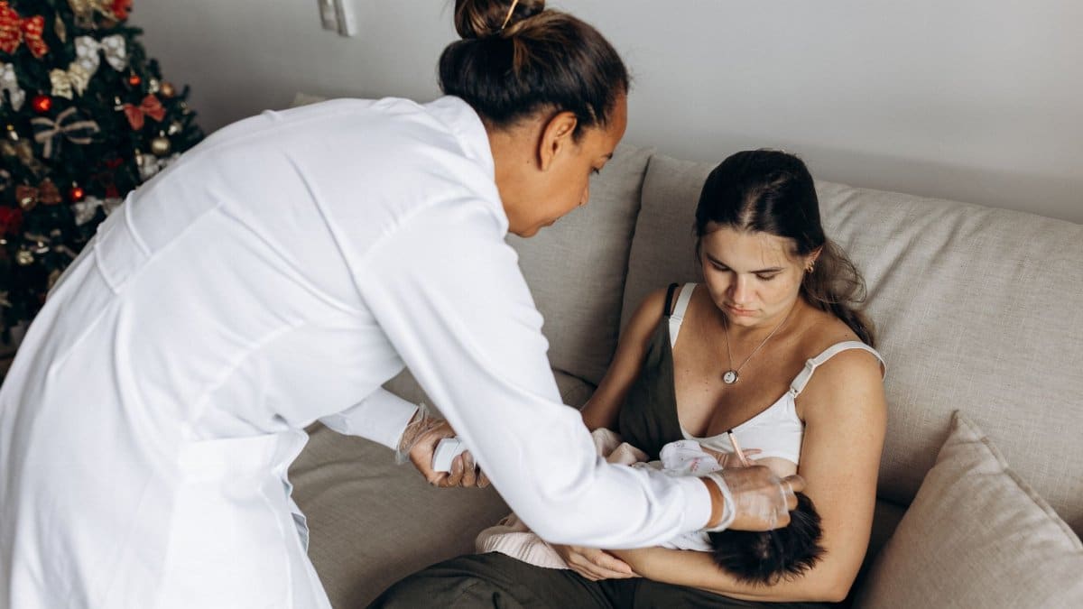 A healthcare professional helps a new mother care for her baby on a sofa during a home visit.