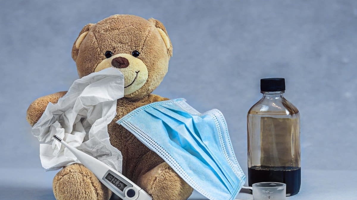 A teddy bear with cough syrup, a mask, thermometer, and tissue highlights an illness concept.