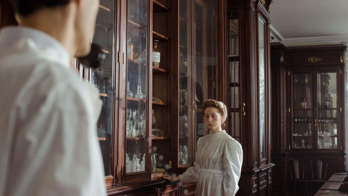 Vintage interior with woman in white dress examining wooden cabinets, evoking a historical apothecary setting.