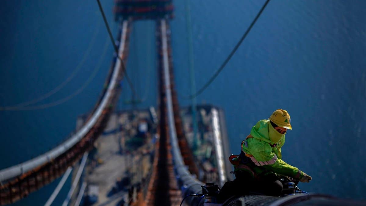 A construction worker fixes cables on a bridge high above the water, showcasing engineering and safety.
