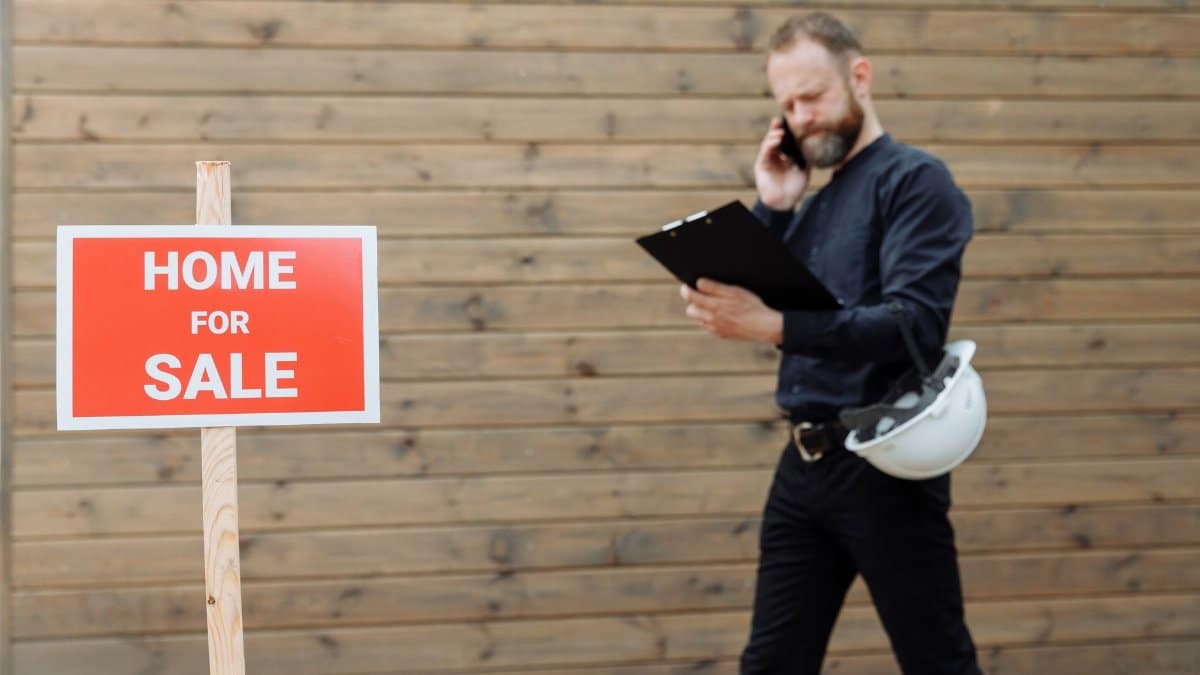 Real estate agent on phone with 'Home for Sale' sign outside a property.