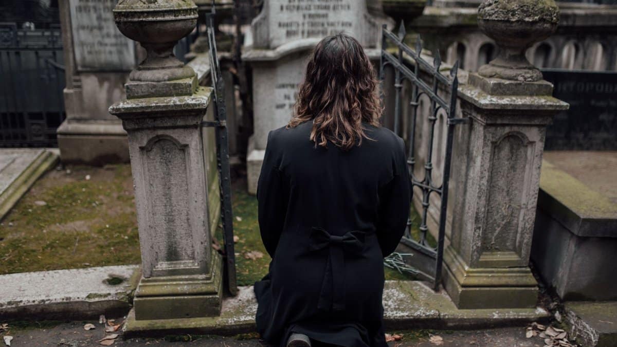 A woman in a black coat kneels at a grave in a gloomy outdoor cemetery.