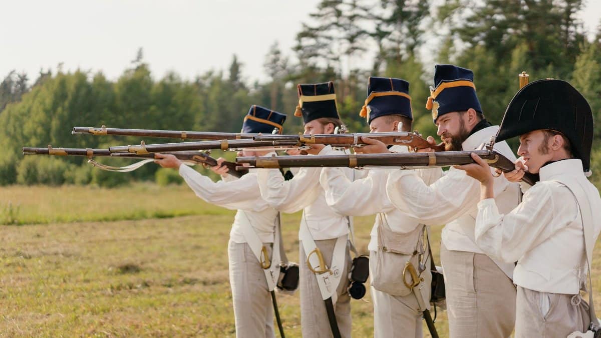 Soldiers in vintage uniforms aiming rifles during a historical battle reenactment outdoors.