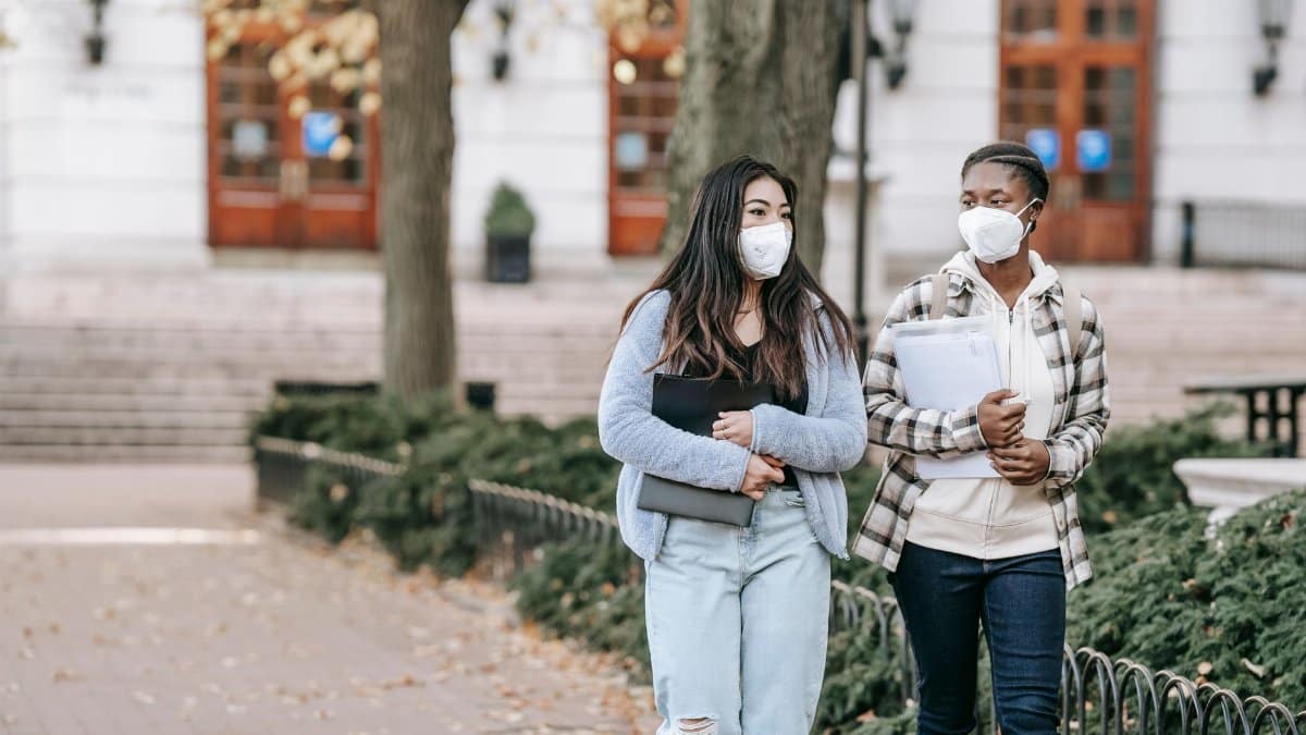 Calm young multiracial female students with folders wearing casual clothes and face masks strolling together on campus sidewalk