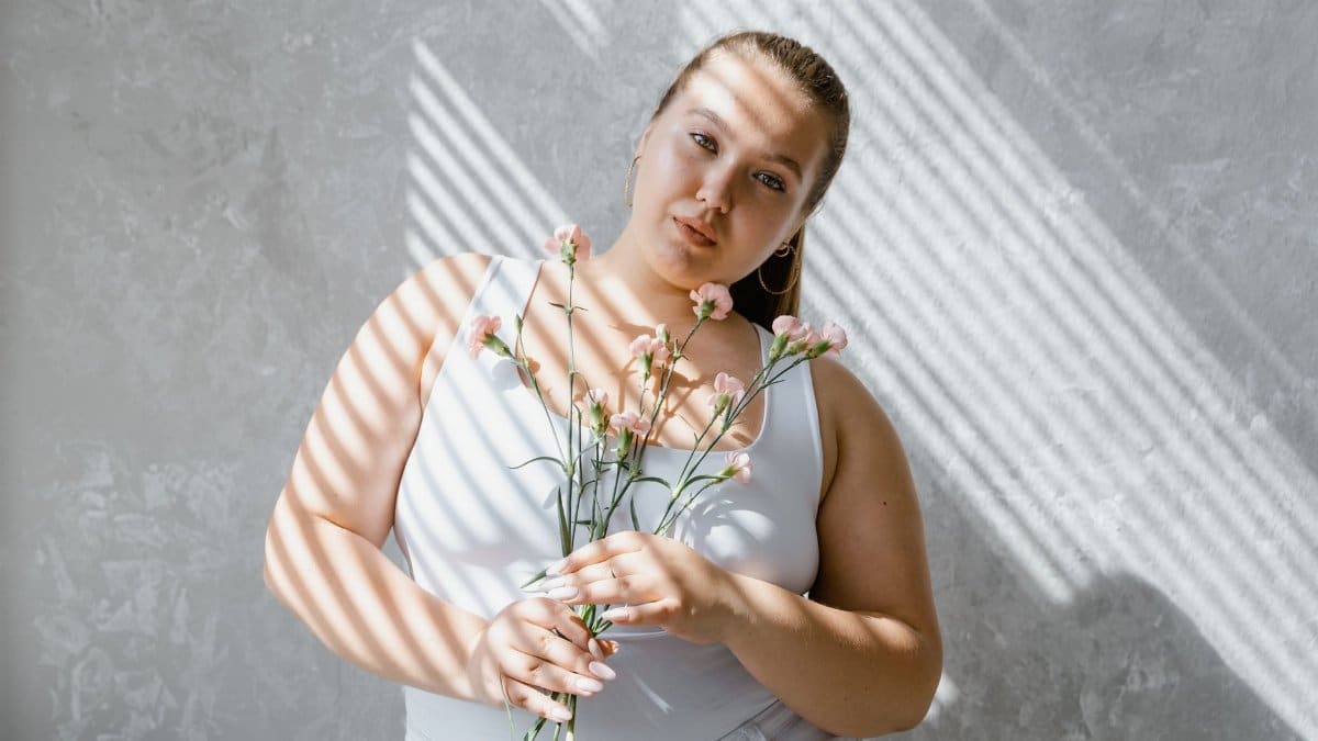 Plus-size woman in tank top holding flowers with sunlight filtering through blinds.