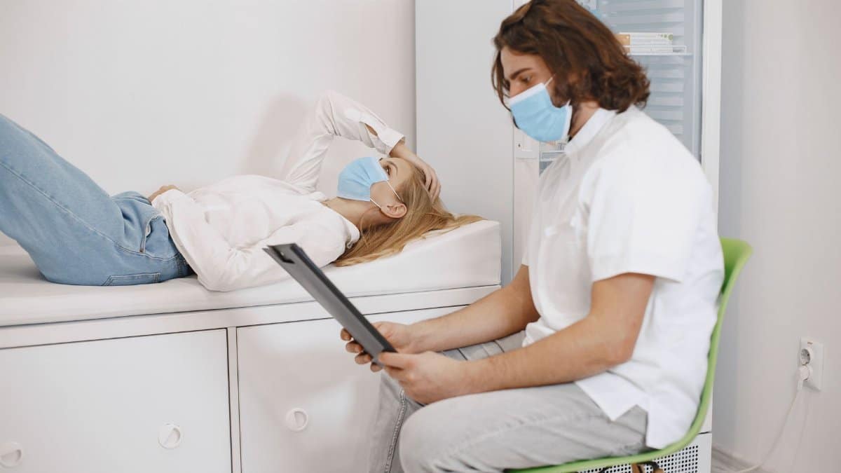 Doctor consulting patient wearing face masks in a medical examination room, discussing healthcare plan.