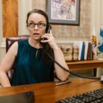Businesswoman in office attire talking on a landline phone at her desk.
