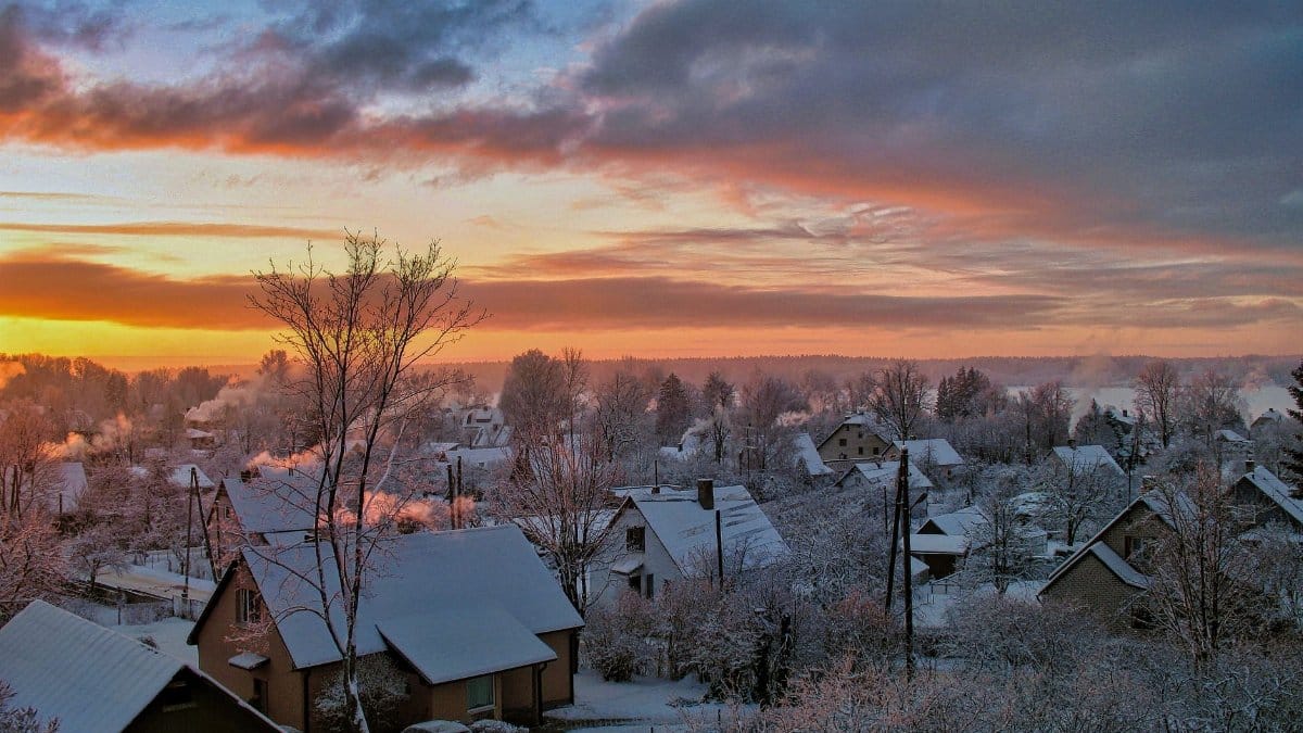 A serene snowy village landscape at sunrise, with houses under snow and a colorful sky.