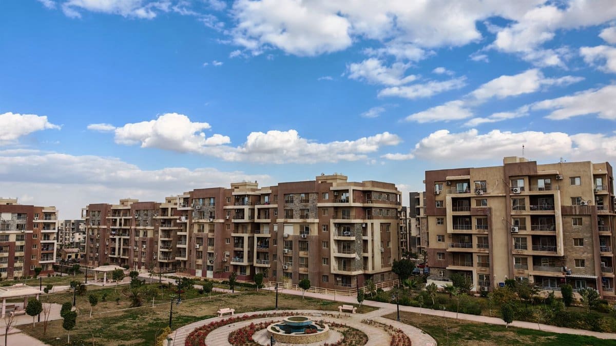 Aerial view of a modern residential complex with a central fountain under a blue sky and clouds.