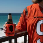 Man in Bengals jersey enjoying beer by the beach on a sunny day.