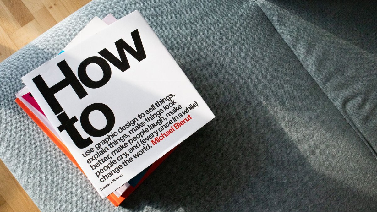 A collection of design books on a couch, showcasing 'How to' by Michael Bierut