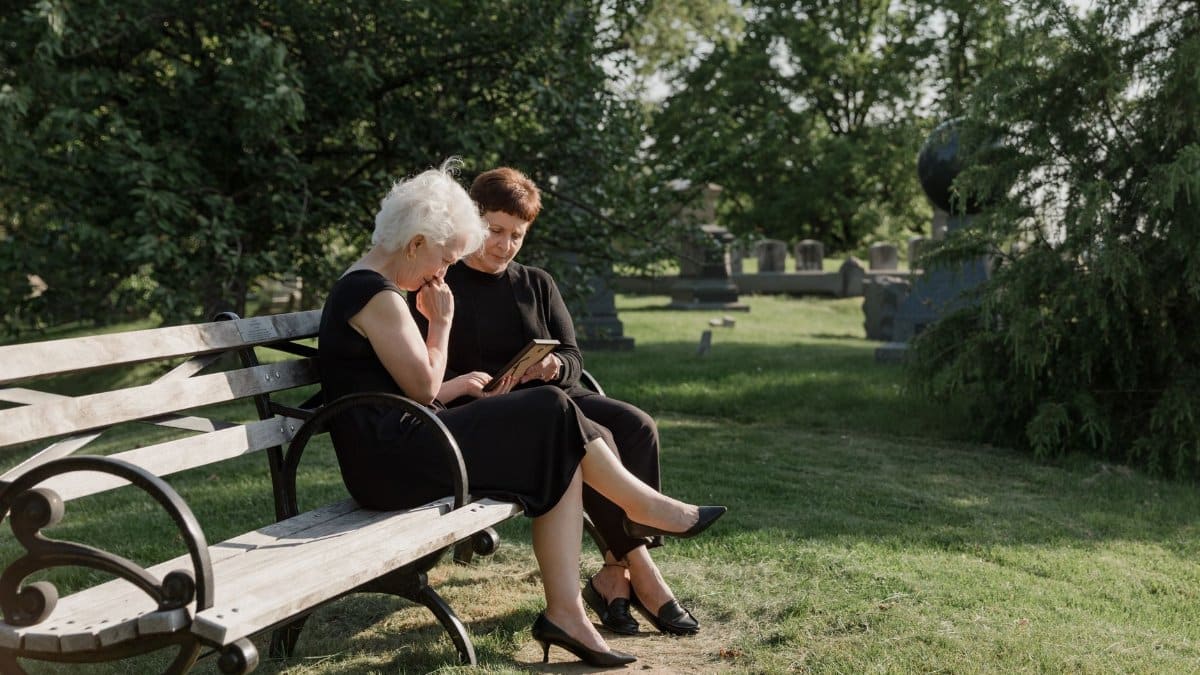 Two women sit on a bench in a peaceful cemetery, reflecting and sharing memories.