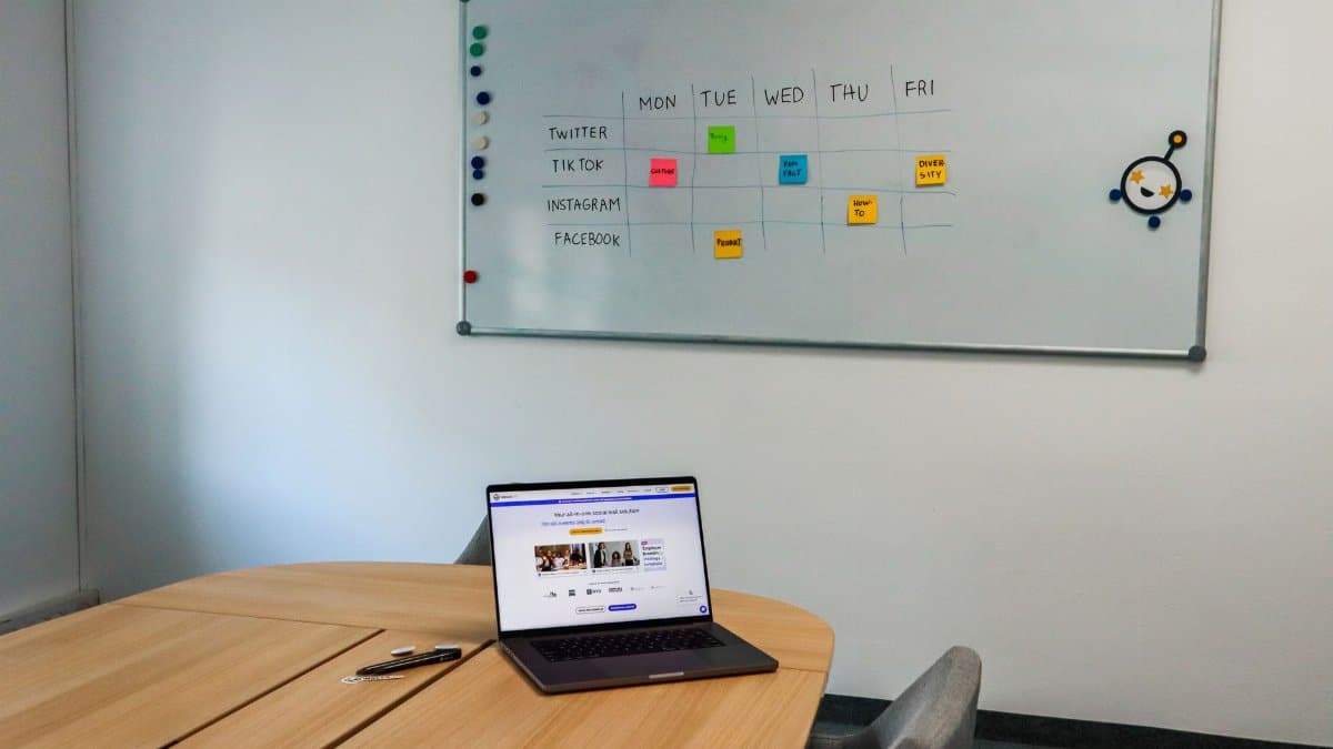 Office setup featuring a conference table, laptop, and organized whiteboard schedule, highlighting a tech-driven workspace.