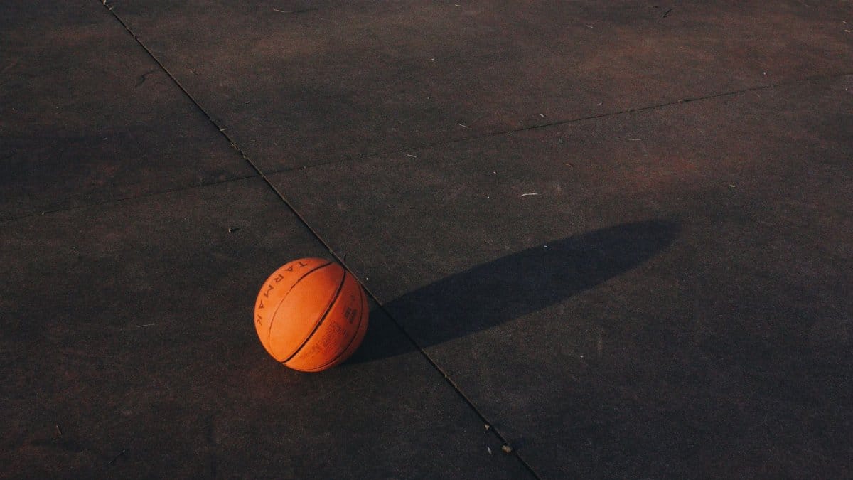 A single basketball casting a shadow on a textured outdoor court surface at dusk.