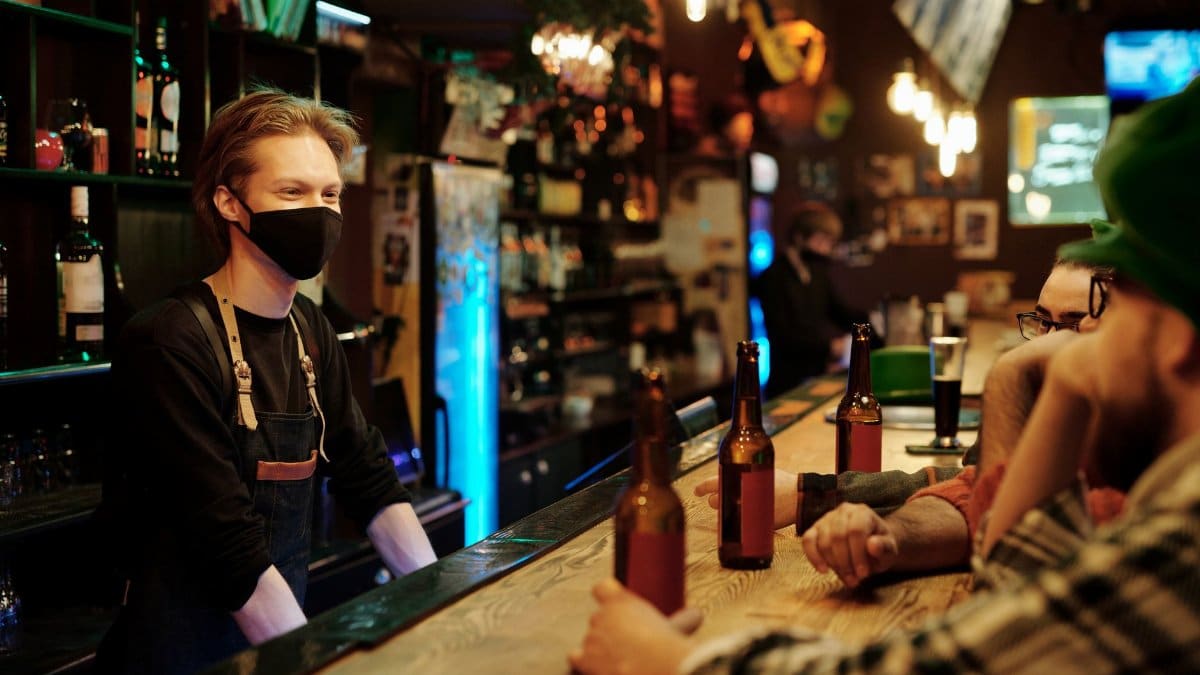 A bartender wearing a mask serves drinks to patrons at a lively bar.