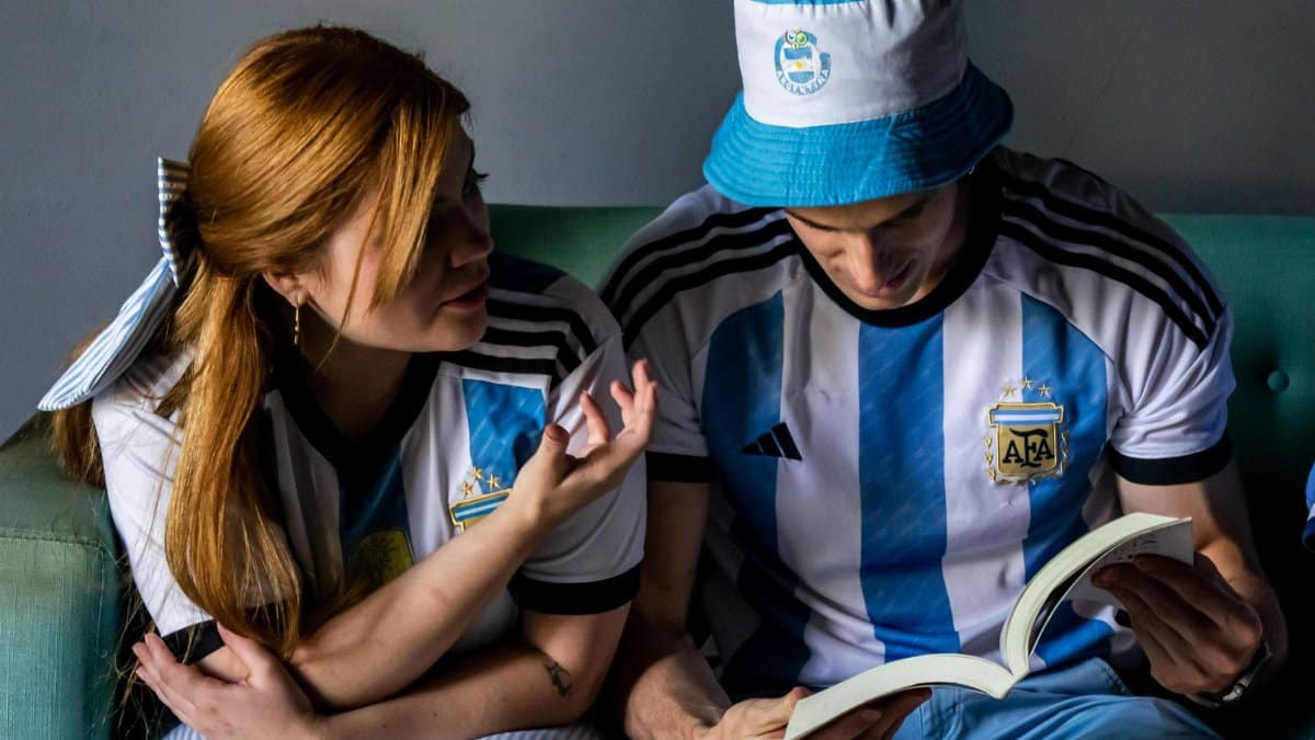 Two people in Argentina soccer jerseys reading a book together on a sofa indoors.