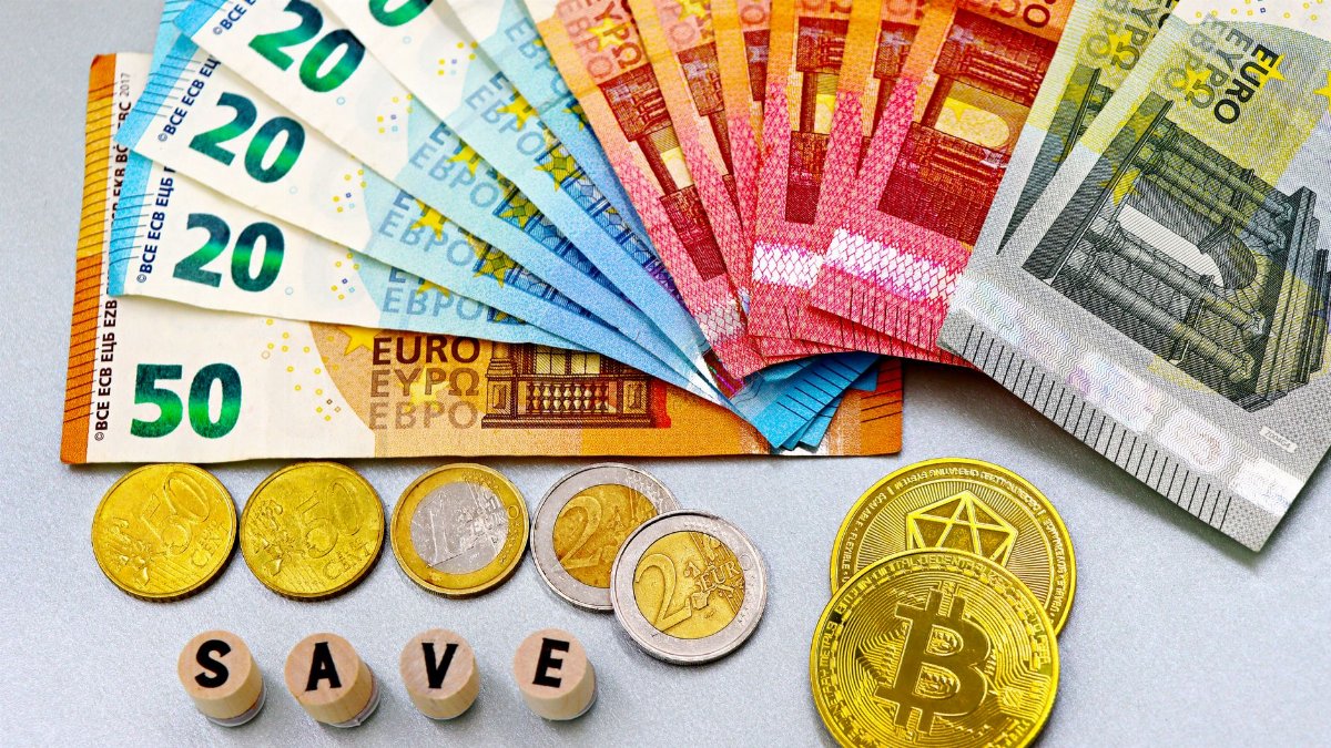 Euro banknotes and cryptocurrency coins arranged on a white surface with blocks spelling 'SAVE'.