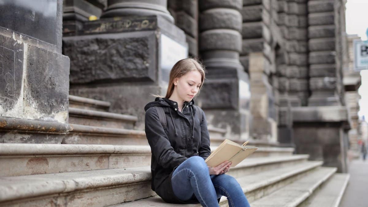Young woman sitting on stone steps of a historic building reading a book outdoors.