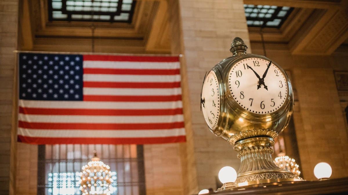 From below of golden old fashioned clock placed in building with ornamental walls against American flag placed in Grand Central Terminal in New York City