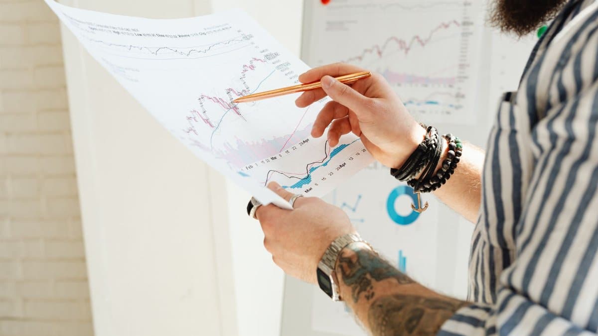 A man analyzing stock market charts with a pen, holding a paper report indoors.