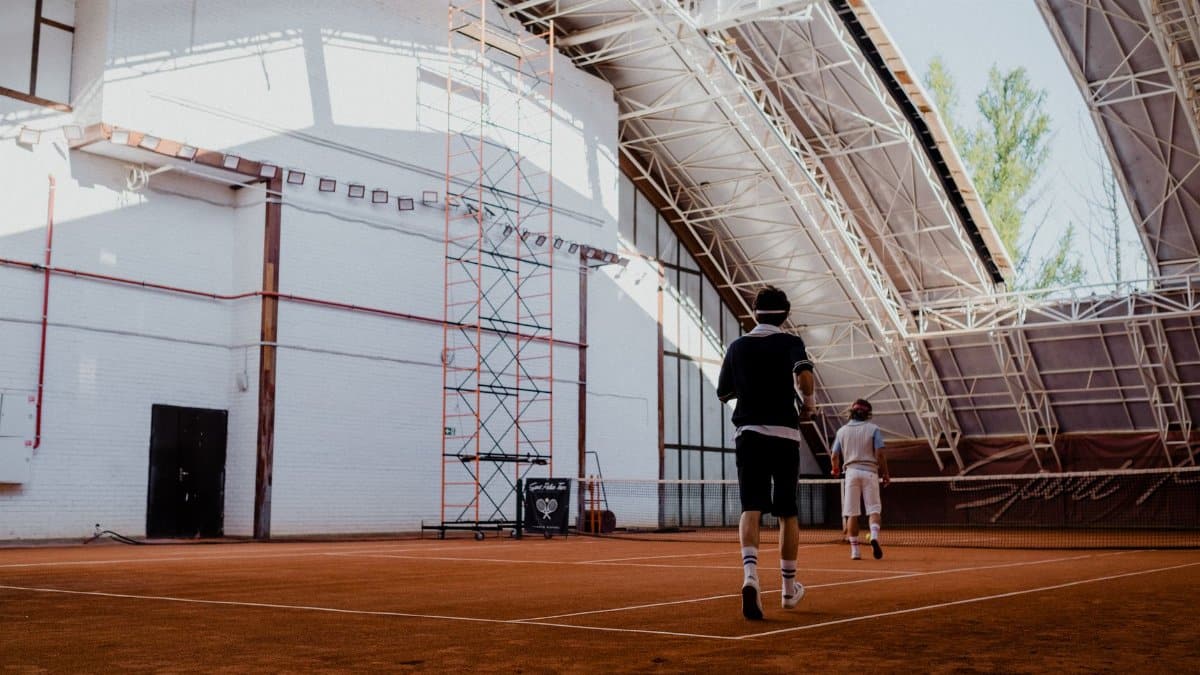 Two players engaging in a tennis match on an indoor clay court with natural lighting.