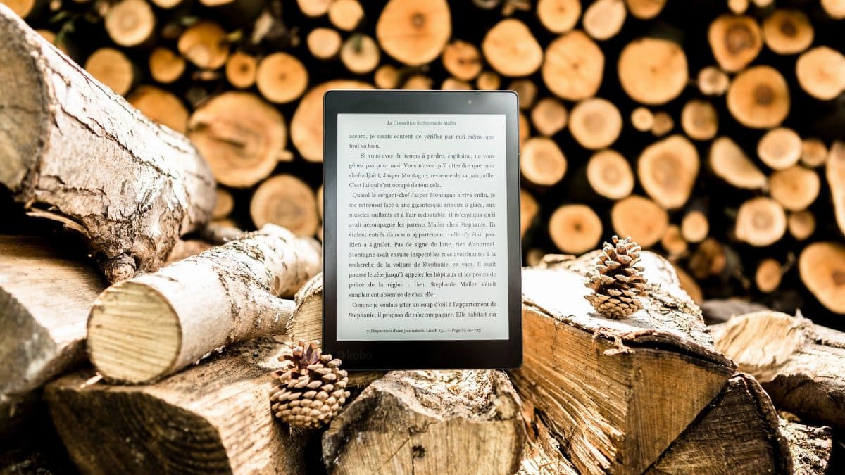 E-reader placed on a pile of wooden logs surrounded by pine cones outdoors.