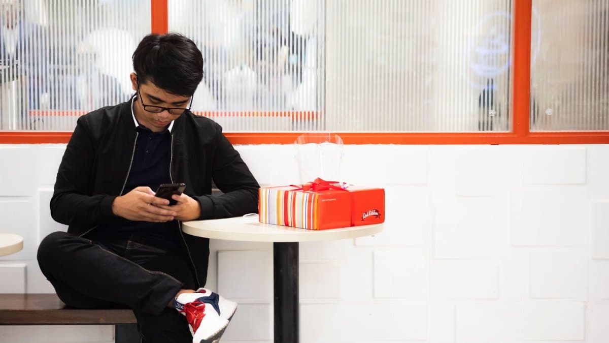 Concentrated young guy in casual clothes sitting on bench near table with present box and window while surfing on smartphone in light cafeteria