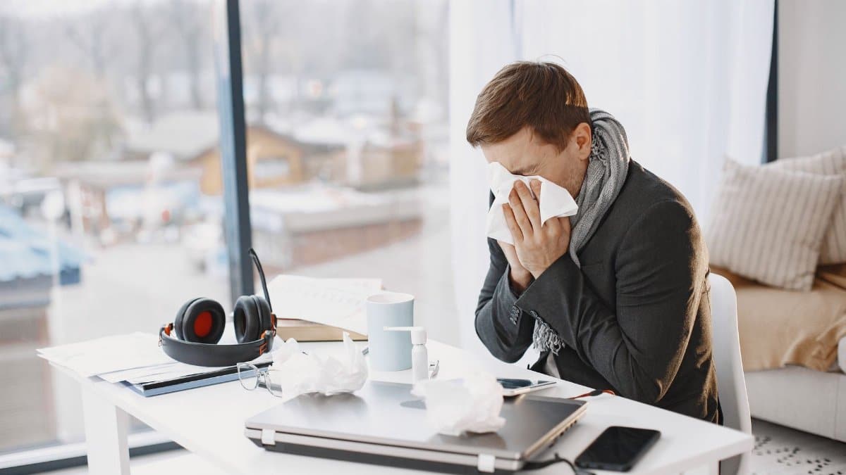 Man sneezing at home desk, indicating remote work during illness.