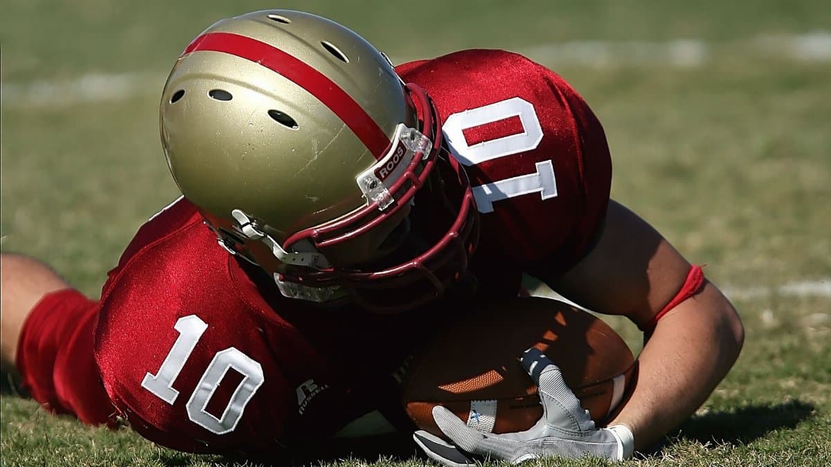 American football player wearing red uniform holding ball on grassy field during day game.