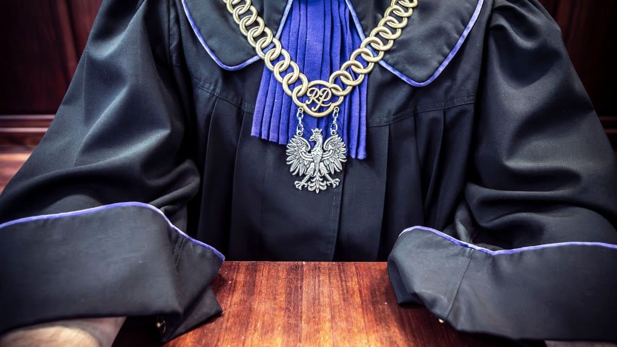 Close-up of a judge wearing a traditional robe with a golden chain medallion, seated at a courtroom table.