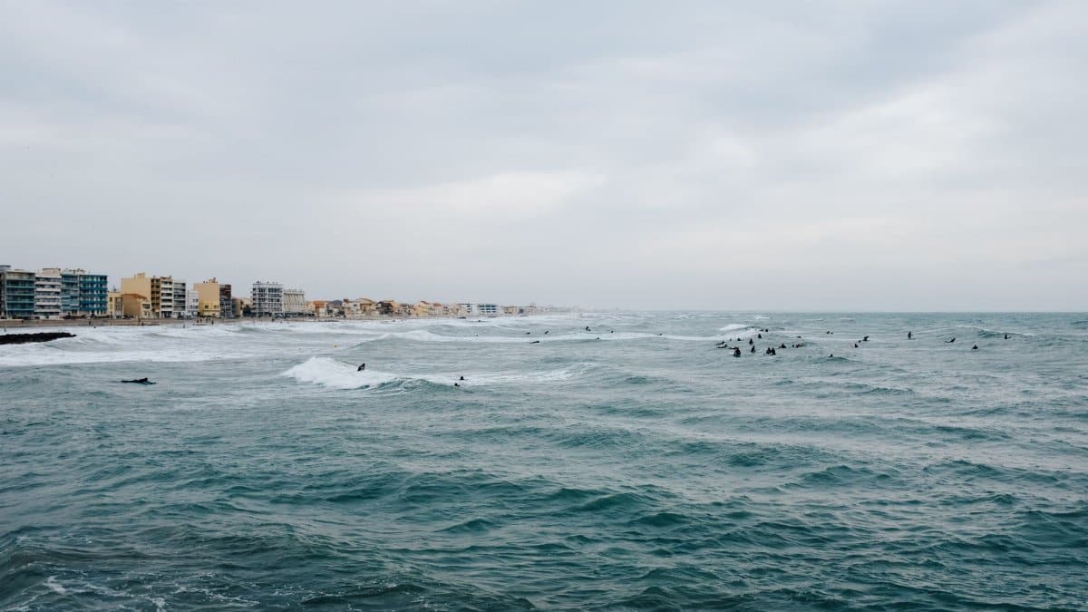 Surfers ride the waves near a coastal city, with buildings lining the shore under a cloudy sky.