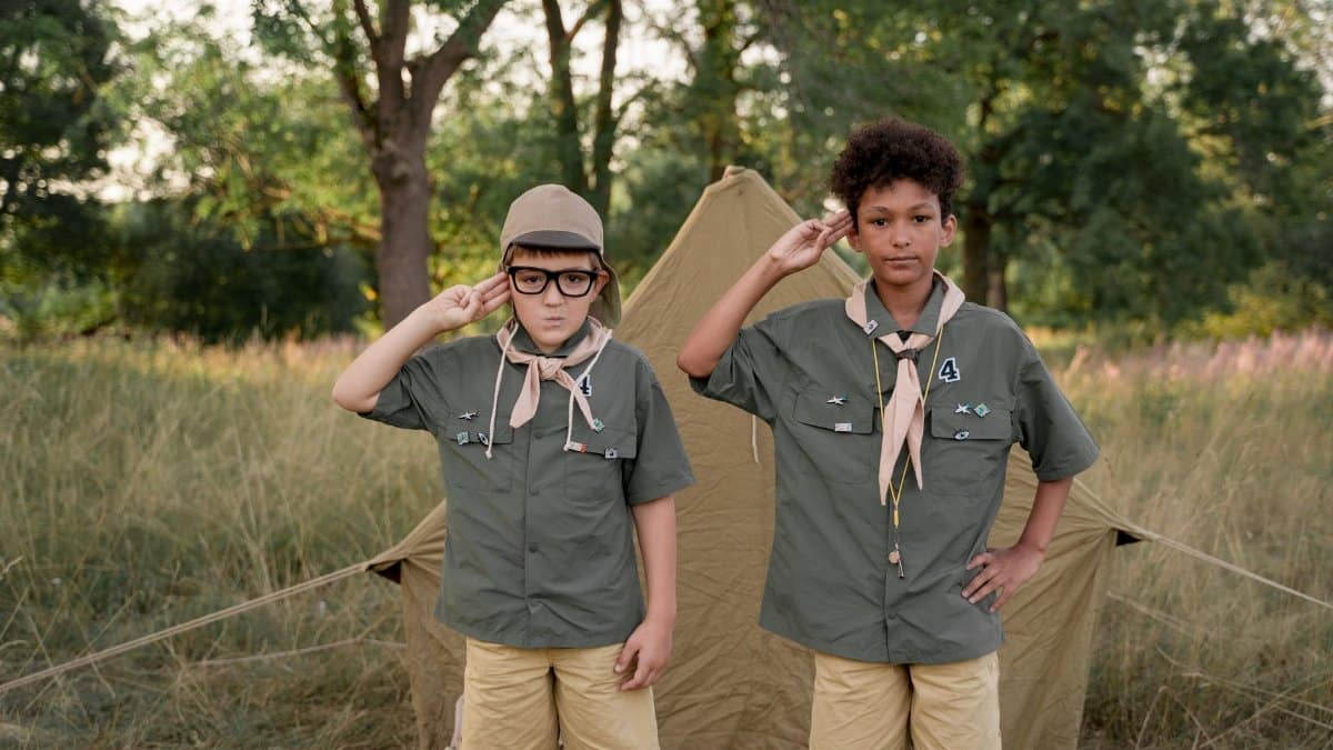 Two boys in scout uniforms saluting in front of a tent during a camp outdoors.