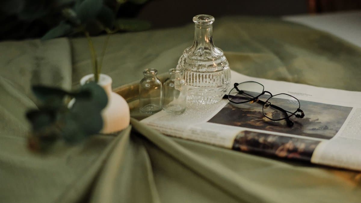 A serene still life featuring glass bottles, eyeglasses, and a newspaper on a draped green fabric.
