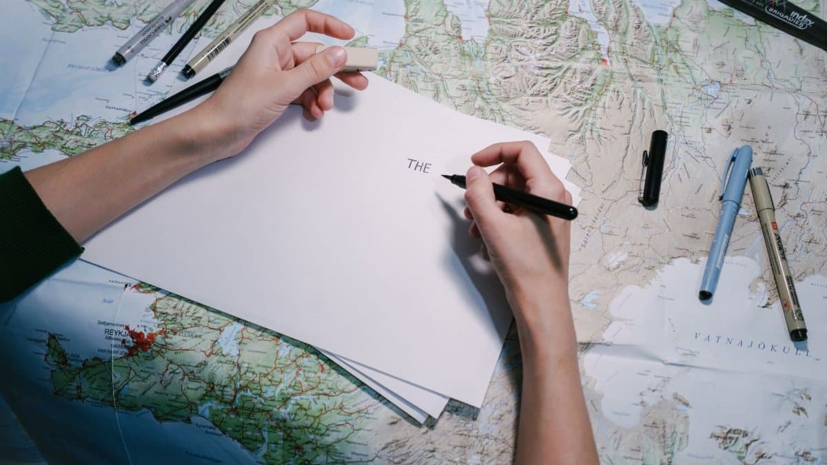 Hands writing on paper over a detailed world map with various pens.