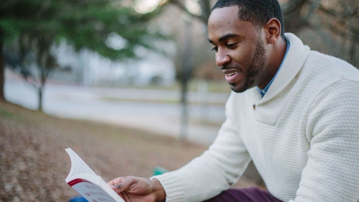 A young man enjoys reading in a serene park setting during the day.