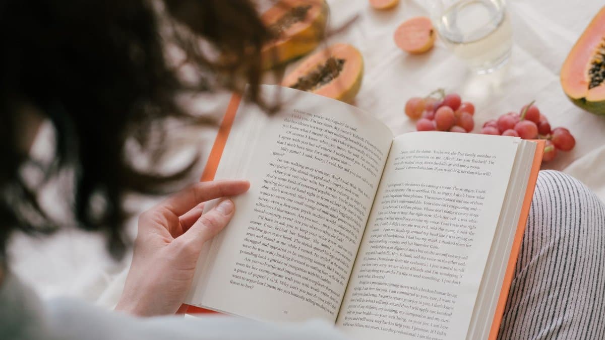 A person enjoys reading a book surrounded by fresh papaya, grapes, and a drink, creating a cozy indoor scene.