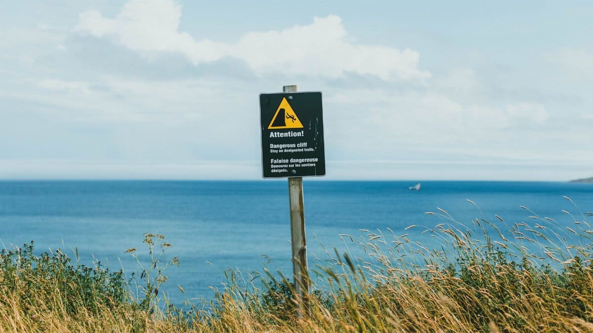 A warning sign on a coastal cliff, with a view of the ocean on a clear day.