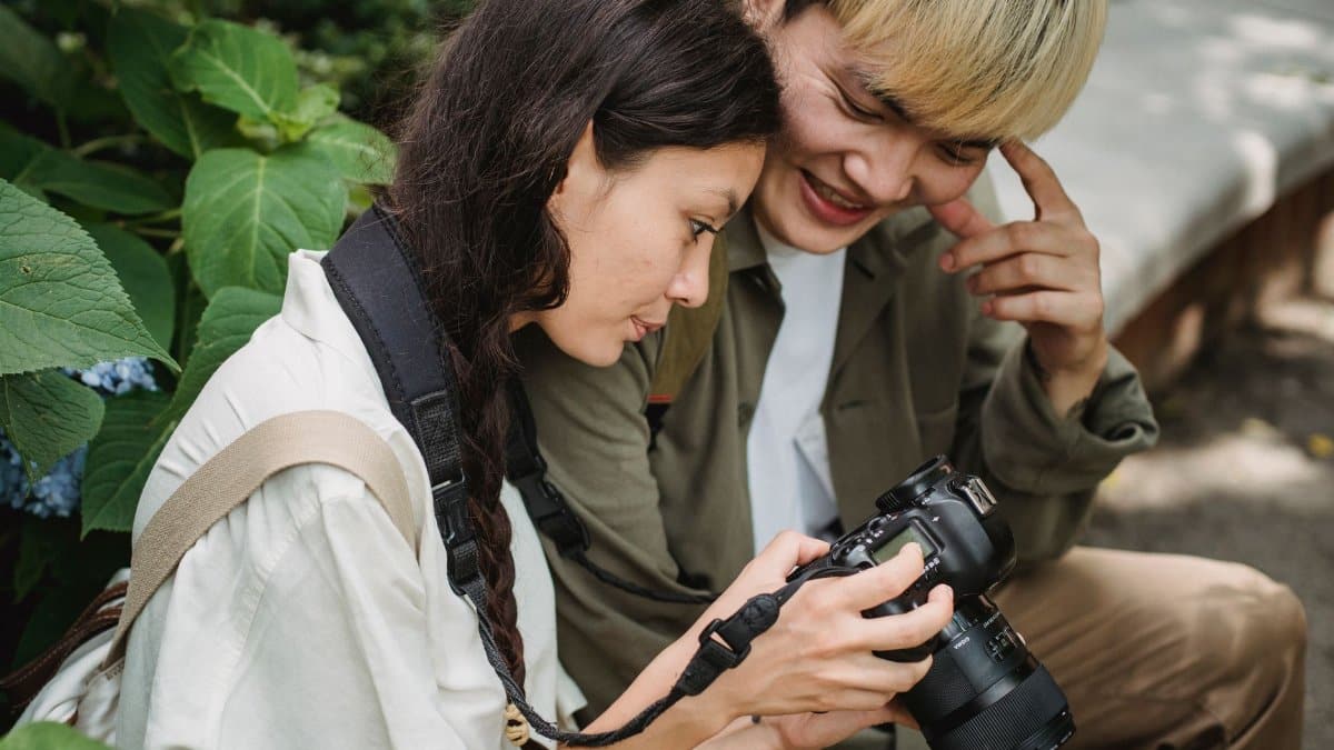 Happy young Asian couple in casual wear looking through photos on professional photo camera while spending warm summer day in garden